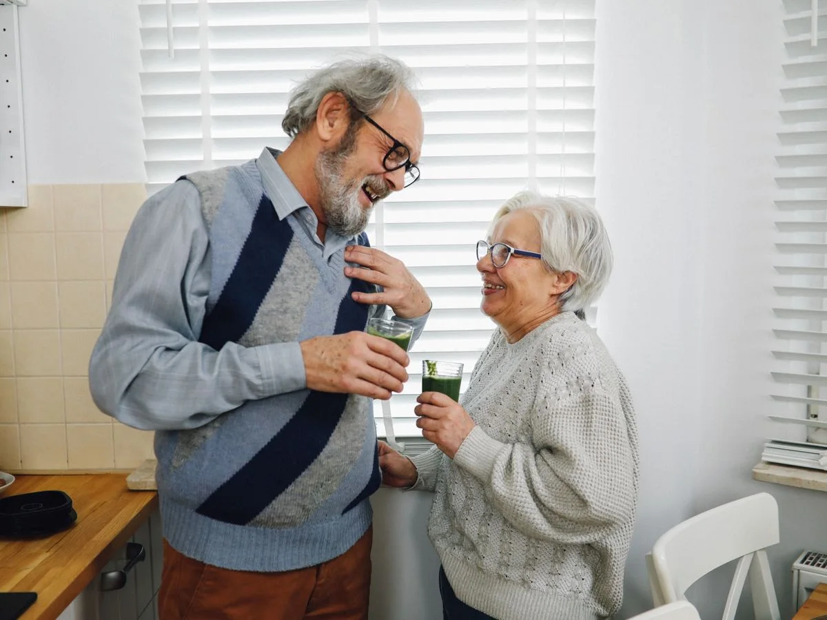 Elderly couple smiling and holding drinks in a cozy kitchen setting.