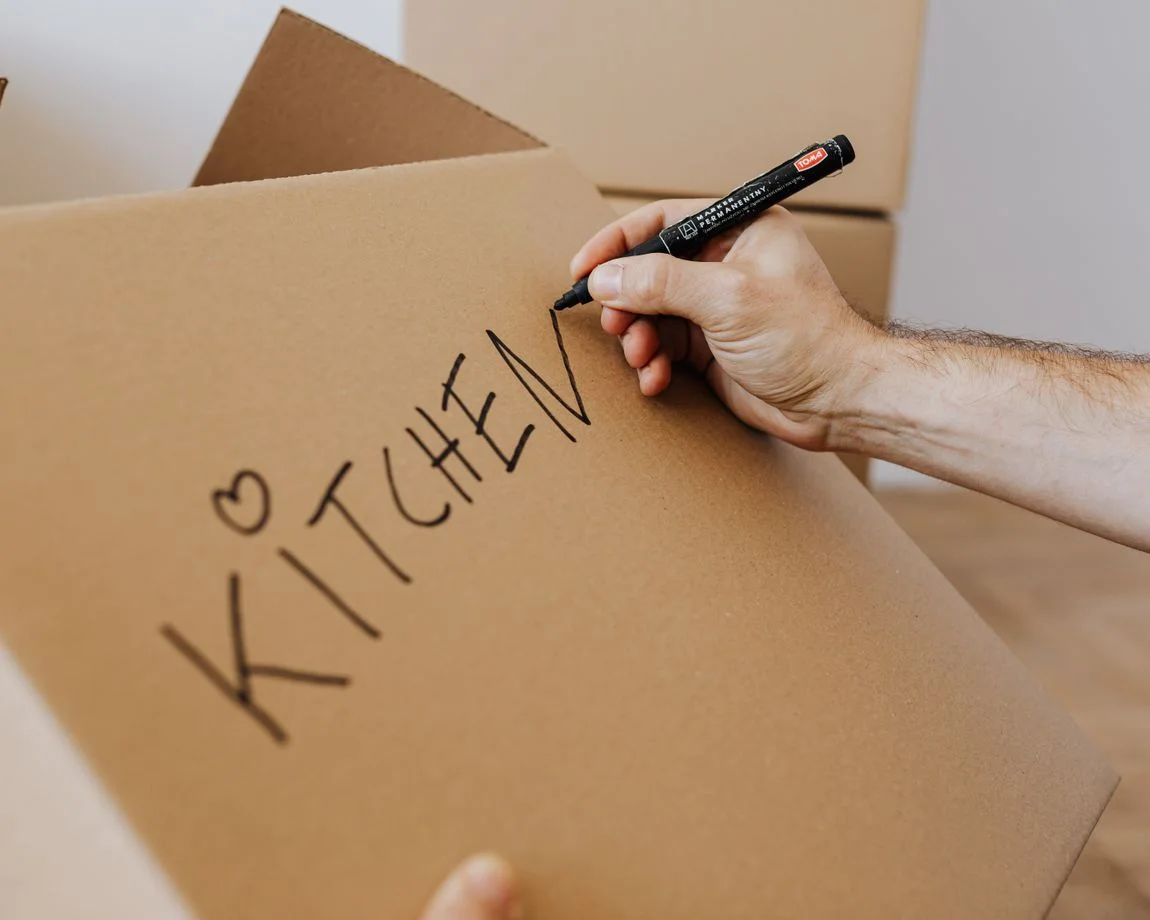 Crop unrecognizable male using marker to write on carton box word kitchen and draw cute heart while packing kitchenware before relocation
