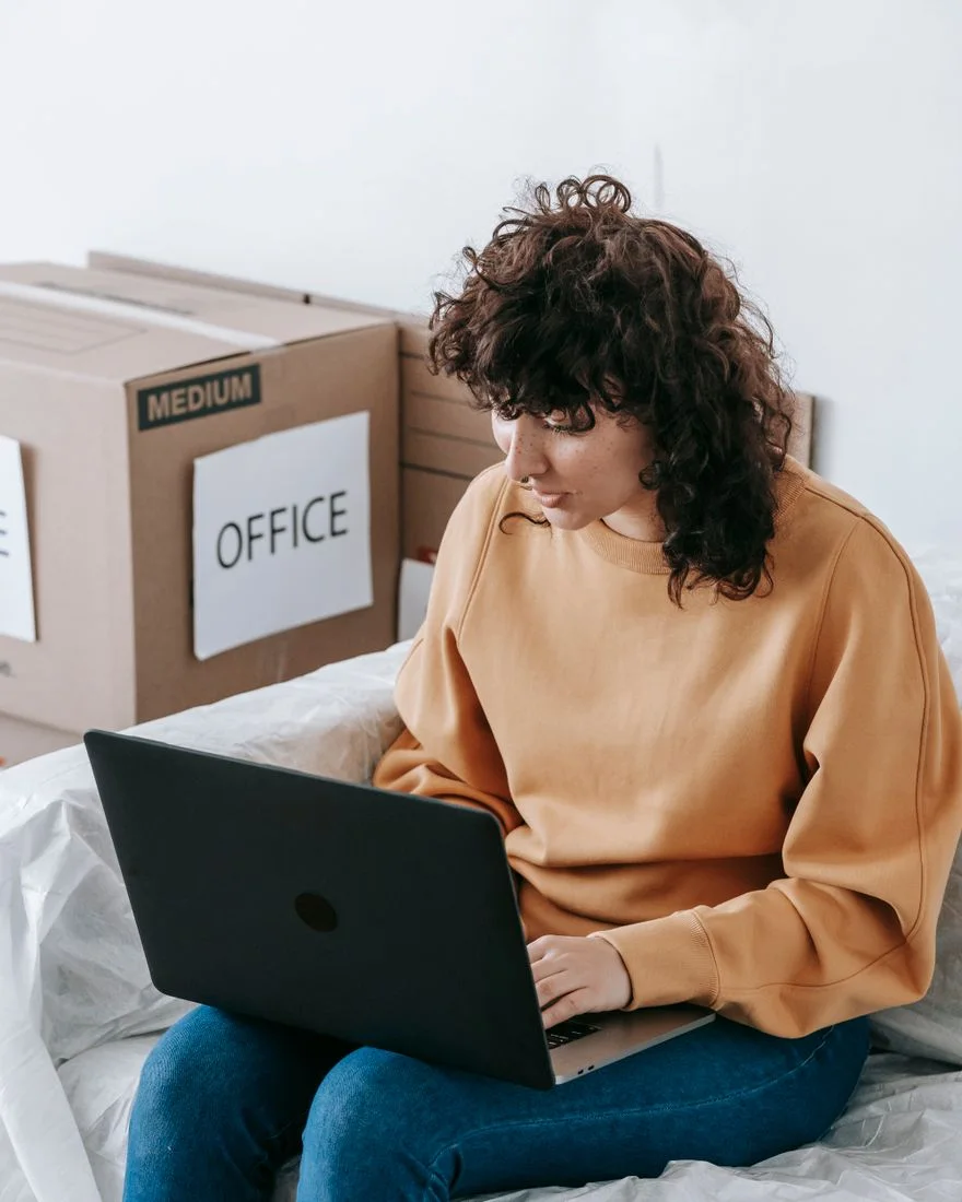 A woman in a casual setting using a laptop on a sofa, surrounded by moving boxes labeled 'OFFICE.'