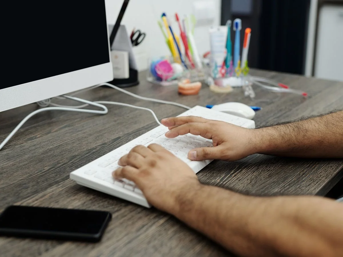 Close-up of hands typing on a white keyboard at a wooden desk in an office setting with accessories.