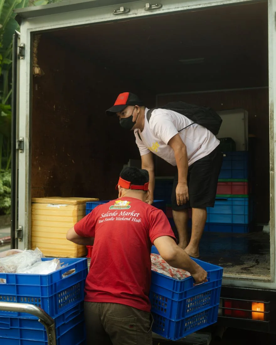 Two men loading blue crates into a delivery truck at a market.