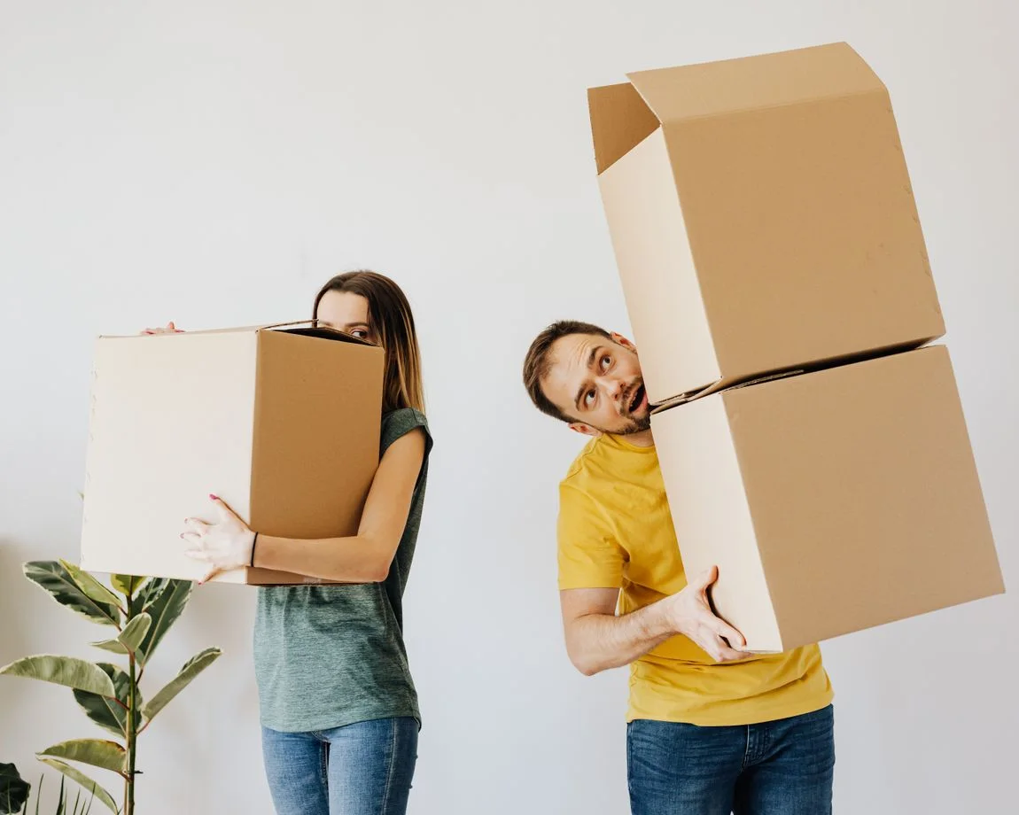 Content couple in casual wear carrying stacked cardboard boxes with belongings while moving into modern apartment together in daylight