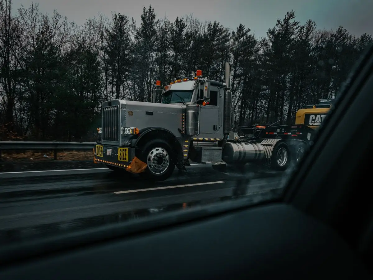 A large truck with an oversize load on a rainy highway, captured through a car window with dark, moody tones.