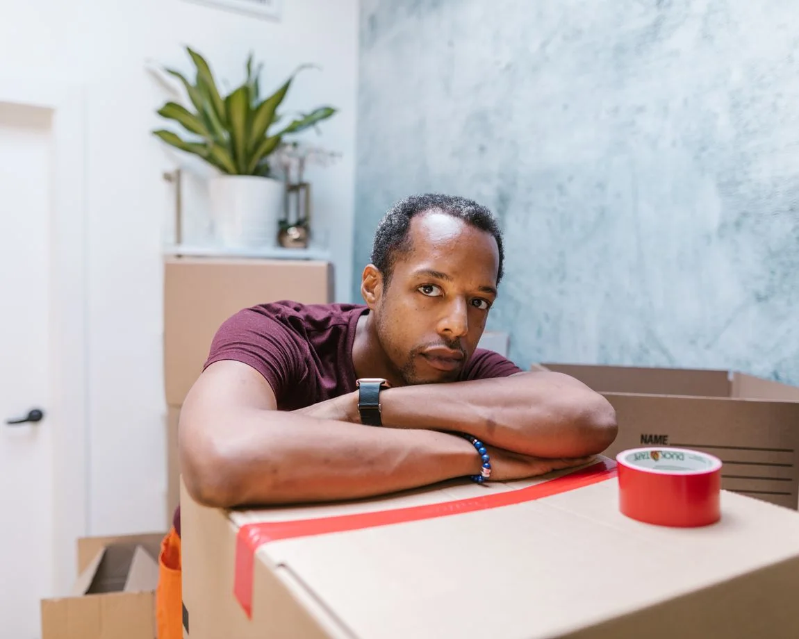 A man resting on cardboard boxes in a room with packing tape visible, suggesting moving or packing activities.