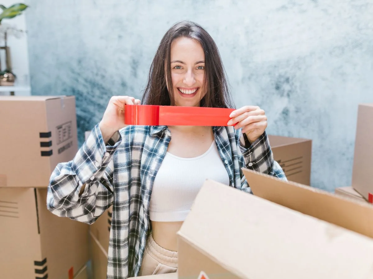 Caucasian woman packing boxes with red tape, smiling in an indoor setting. Perfect for moving and relocation themes.