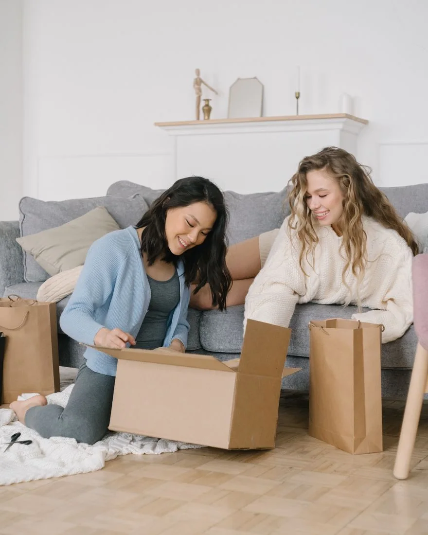 Two women sit on the floor, joyfully unpacking boxes in a cozy living room setting.