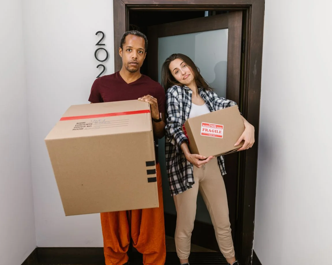 A couple carrying moving boxes indoors, settling into their new apartment.