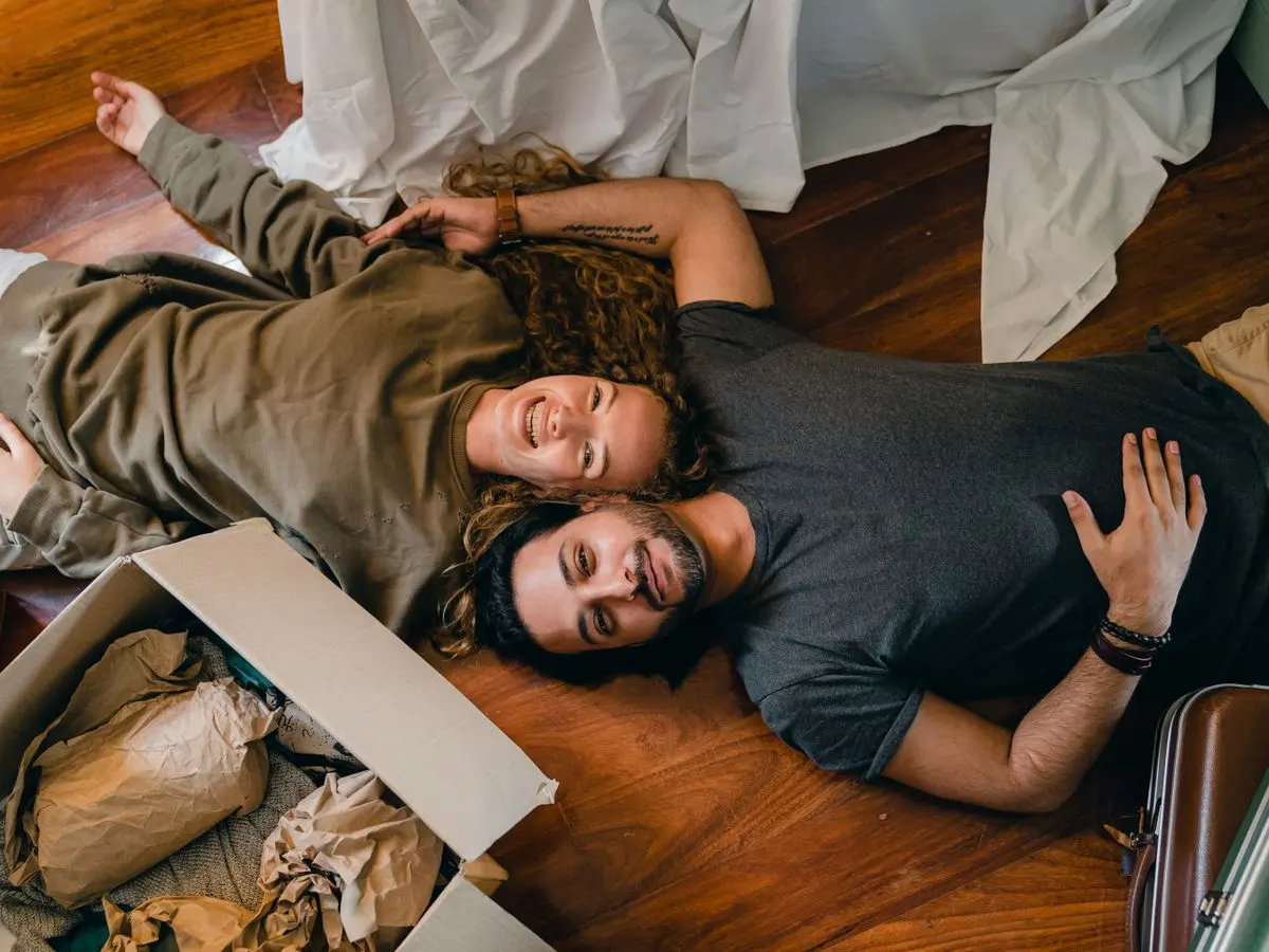 From above of young smiling diverse couple in casual clothing relaxing on floor near carton boxes while unpacking belongings in new contemporary apartment