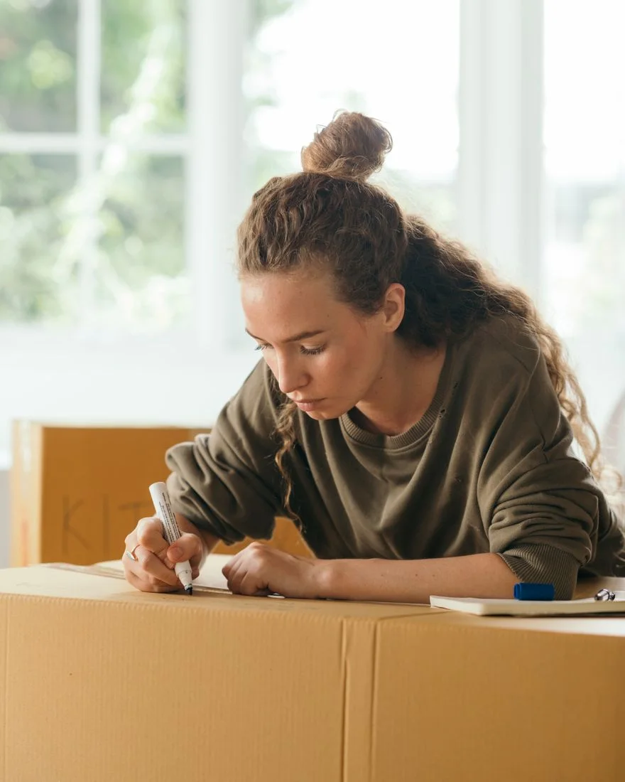 Concentrated long curly haired lady signing with marker carton boxes with packed things for moving at daytime