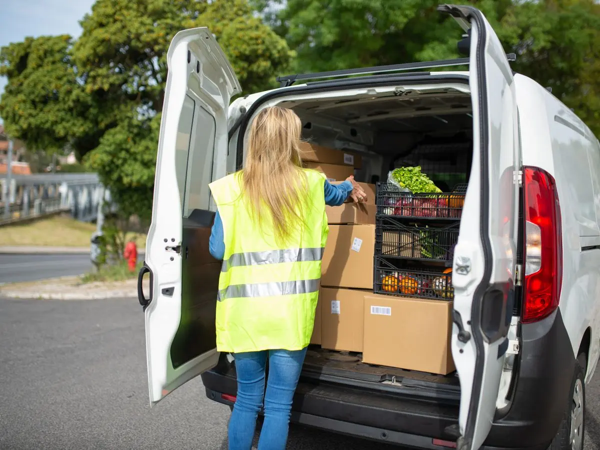 A woman in a reflective vest loads groceries and packages into a delivery van on a sunny day in Portugal.