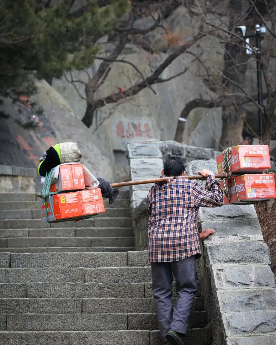 Elderly man carrying boxes on a pole up stone steps, showcasing resilience and tradition.