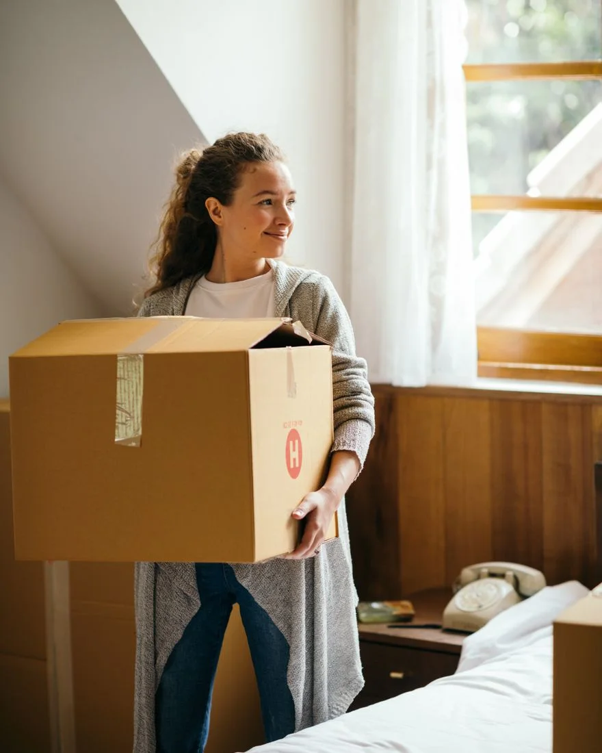 Satisfied female with curly hair smiling while carrying carton box moving out house