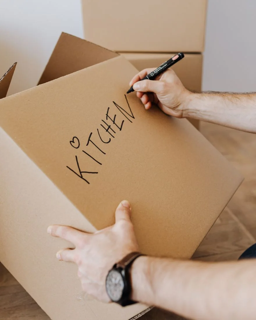 Crop unrecognizable male using black marker to write on carton container word kitchen and draw heart to identify box with packed kitchenware during relocation