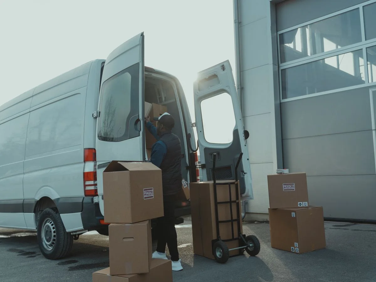 Courier loading cardboard boxes into a delivery van outside a warehouse.