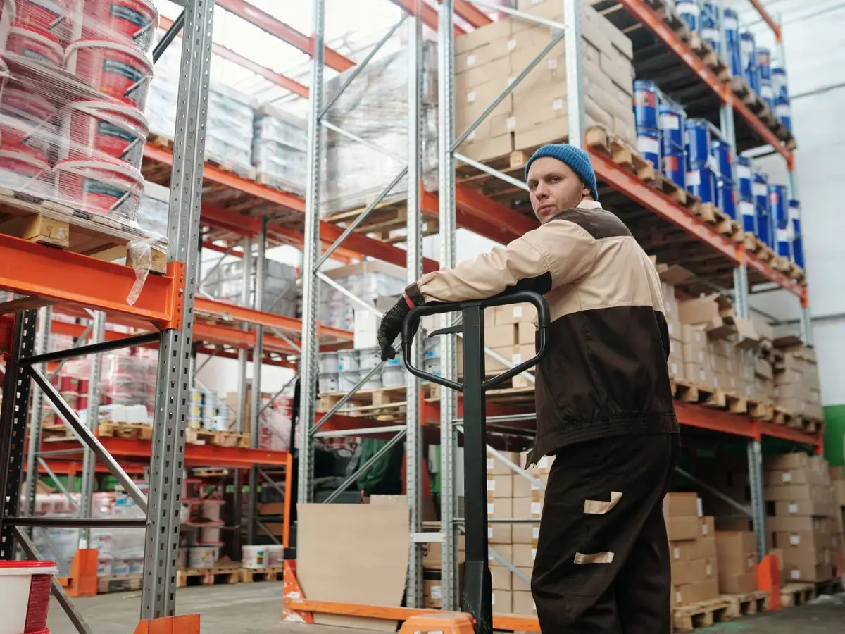 Warehouse worker operating a pallet jack among stacked boxes and metal shelves in an industrial setting.