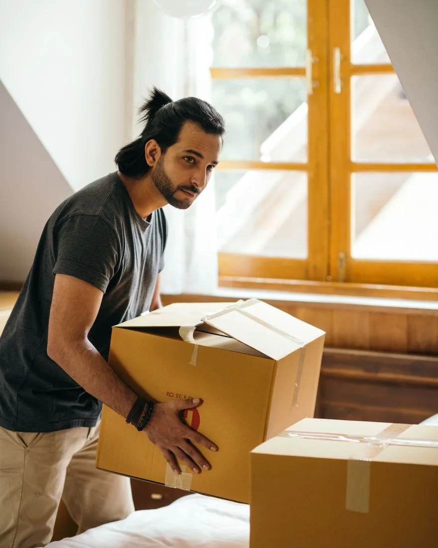 Side view of pensive ethnic male in casual wear and smart watch looking away while taking cardboard box from bed and standing near window after moving into new house