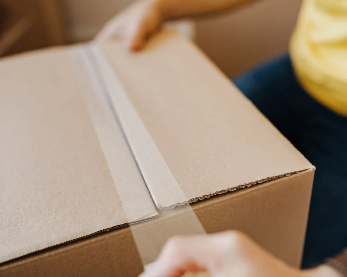 Crop anonymous courier in yellow shirt packing purchase and sealing carton box with tape before deliver