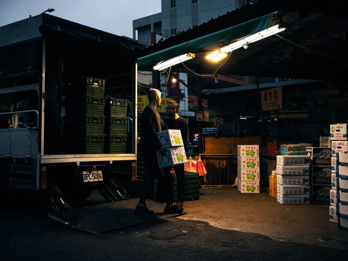 Workers unloading boxes from a delivery truck at an urban market during dusk.